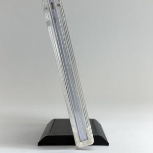 May include: A clear, rectangular acrylic display stand with a black base. The stand is angled, showcasing a vertical, ribbed design. The base is a solid, matte black, providing a stable foundation for the clear display.