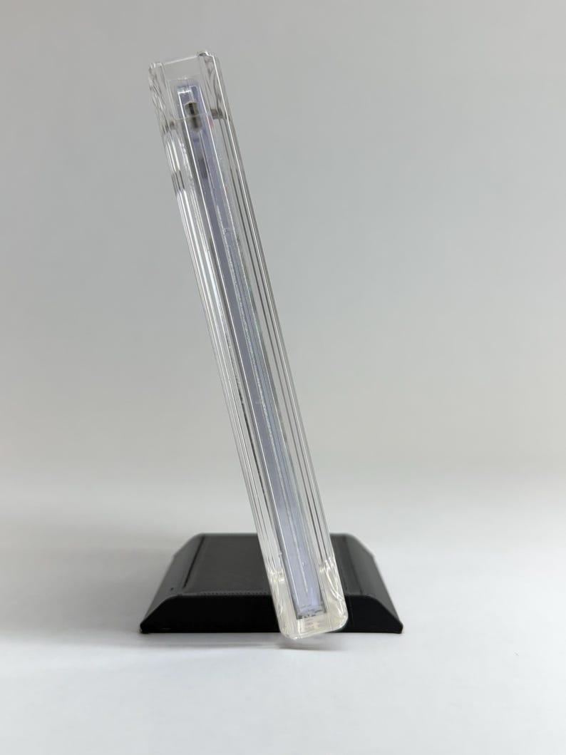 May include: A clear, rectangular acrylic display stand with a black base. The stand is angled, showcasing a vertical, ribbed design. The base is a solid, matte black, providing a stable foundation for the clear display.