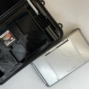 Puede incluir: Una funda rígida negra con un interior gris para una consola de juegos Nintendo DS. La funda tiene ranuras para varios cartuchos de juegos. Una consola Nintendo DS Lite plateada está al lado de la funda. La consola tiene un rasguño en la parte superior.