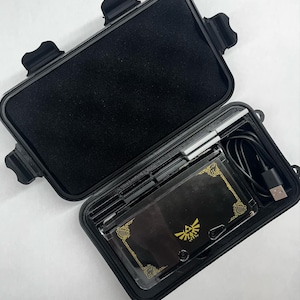 May include: A black hard case with a zippered closure, open to reveal a black Nintendo 3DS console with a gold Triforce design, a stylus, and a charging cable.
