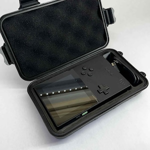 K&ouml;nnte beinhalten: Schwarze Handheld-Spielkonsole mit Bildschirm, Tasten und Steuerkreuz in einem offenen schwarzen Schutzgeh&auml;use. Das Geh&auml;use hat eine Schaumstoffauskleidung und sichere Verschl&uuml;sse. Ein Ladekabel ist ebenfalls sichtbar.