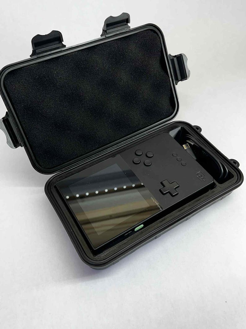 K&ouml;nnte beinhalten: Schwarze Handheld-Spielkonsole mit Bildschirm, Tasten und Steuerkreuz in einem offenen schwarzen Schutzgeh&auml;use. Das Geh&auml;use hat eine Schaumstoffauskleidung und sichere Verschl&uuml;sse. Ein Ladekabel ist ebenfalls sichtbar.