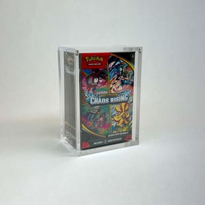 May include: A clear acrylic display case containing a Pokémon Trading Card Game Chaos Rising booster bundle. The box features colorful character illustrations, the Pokémon logo, and text stating it includes 6 booster packs and is rated for ages 6+.