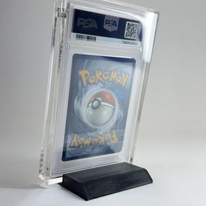 May include: A graded Pok&eacute;mon trading card in a clear plastic case, displayed on a black stand. The card features the Pok&eacute;mon logo and a Pok&eacute; Ball design. The case has a PSA label with a barcode and serial number.