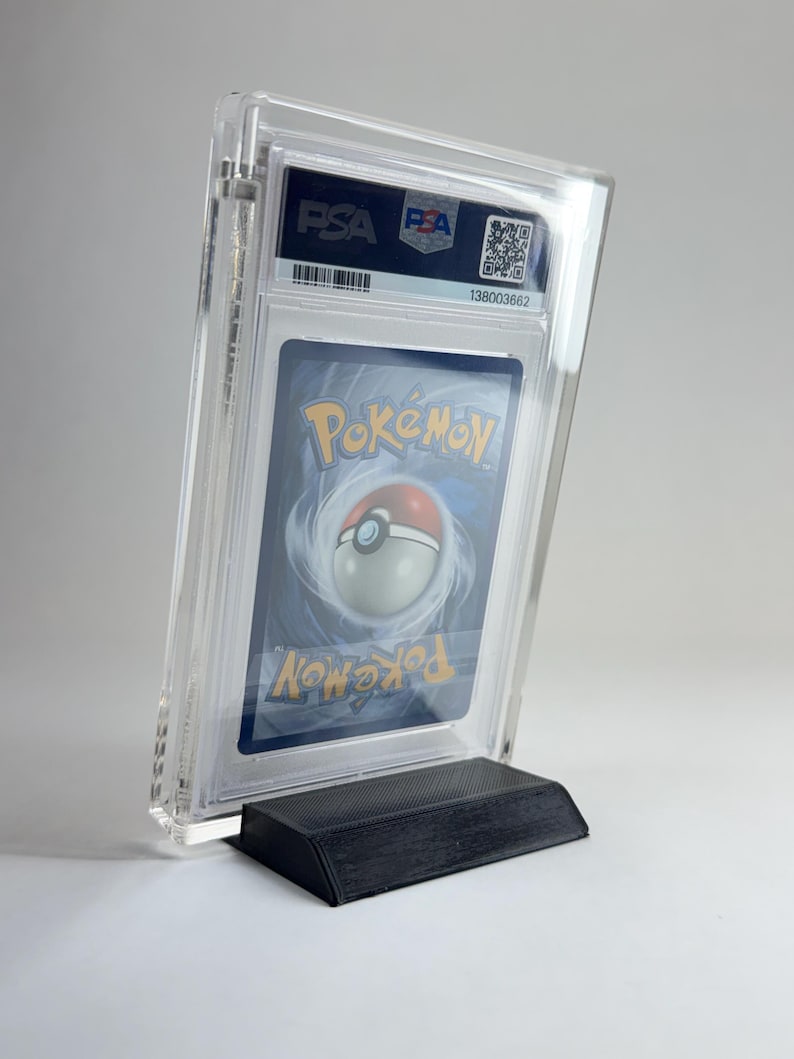 May include: A graded Pok&eacute;mon trading card in a clear plastic case, displayed on a black stand. The card features the Pok&eacute;mon logo and a Pok&eacute; Ball design. The case has a PSA label with a barcode and serial number.