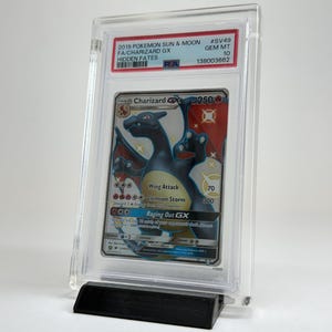 May include: A graded Charizard GX trading card from the 2019 Pok&eacute;mon Sun & Moon Hidden Fates set. The card is encased in a clear protective holder and displayed on a black stand. The card features a black and orange Charizard illustration.