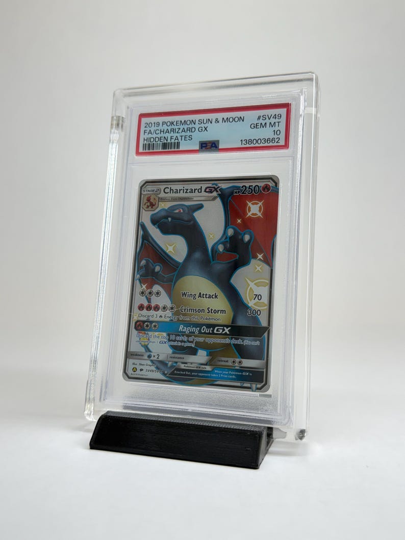 May include: A graded Charizard GX trading card from the 2019 Pok&eacute;mon Sun & Moon Hidden Fates set. The card is encased in a clear protective holder and displayed on a black stand. The card features a black and orange Charizard illustration.
