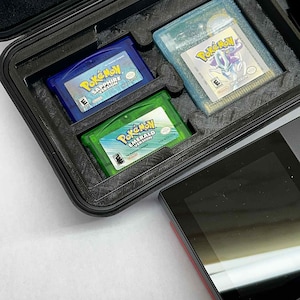 K&ouml;nnte beinhalten: Ein schwarzer Koffer mit drei Game Boy Advance-Cartridges: "Pok&eacute;mon Saphir Version" in Blau, "Pok&eacute;mon Smaragd Version" in Gr&uuml;n und "Pok&eacute;mon" in einem hellblauen Geh&auml;use. Ein schwarzes Handheld-Ger&auml;t befindet sich neben dem Koffer.