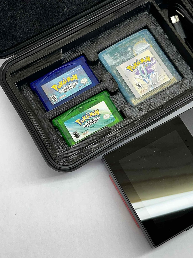 K&ouml;nnte beinhalten: Ein schwarzer Koffer mit drei Game Boy Advance-Cartridges: "Pok&eacute;mon Saphir Version" in Blau, "Pok&eacute;mon Smaragd Version" in Gr&uuml;n und "Pok&eacute;mon" in einem hellblauen Geh&auml;use. Ein schwarzes Handheld-Ger&auml;t befindet sich neben dem Koffer.