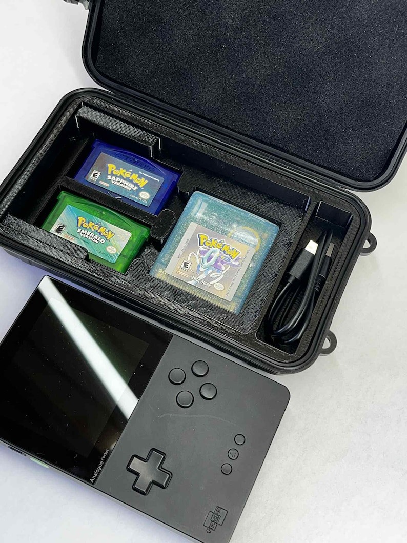 K&ouml;nnte beinhalten: Eine schwarze Handheld-Spielkonsole mit Bildschirm und Steuertasten, neben einem schwarzen Koffer mit drei Pok&eacute;mon-Spielkassetten und einem Ladekabel. Die Kassetten sind blau, gr&uuml;n und transparent.