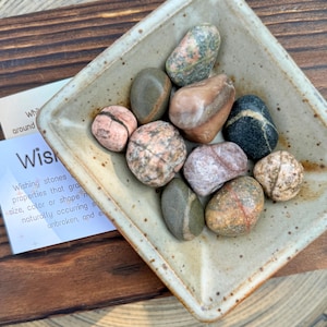 May include: A small, square, light brown ceramic dish with a speckled glaze holds a collection of smooth, colorful stones. The stones are a mix of pink, brown, black, and green with white streaks.