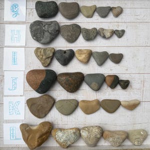 May include: A collection of heart-shaped stones arranged in six horizontal rows, labeled with the letters G, H, I, J, K, and L on small white squares. The stones vary in color, including shades of gray, brown, tan, and black, with diverse textures.