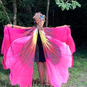 May include: A brown top hat adorned with colorful butterfly decorations and a large, flowing fabric cape featuring a vibrant pink, yellow, and black floral petal pattern that extends outward like wings, worn over a simple black sleeveless top and skirt.