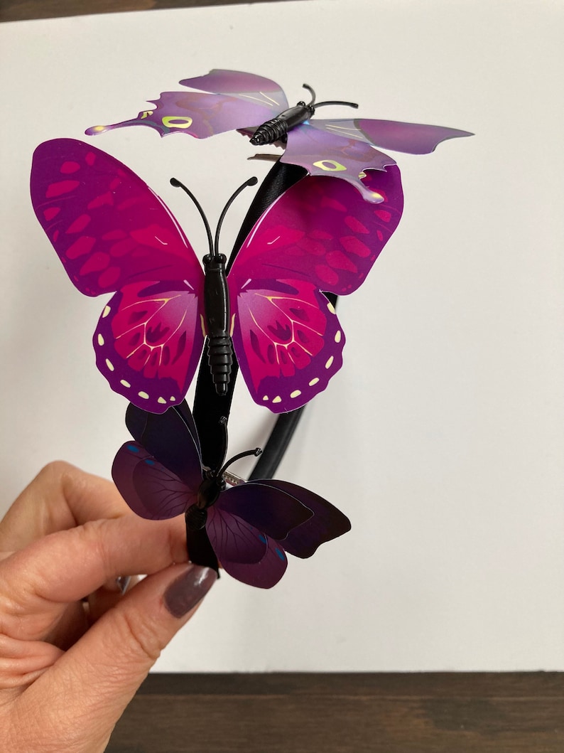 May include: A black headband with three purple butterflies attached. The butterflies have iridescent wings with a dark purple and light purple color gradient.