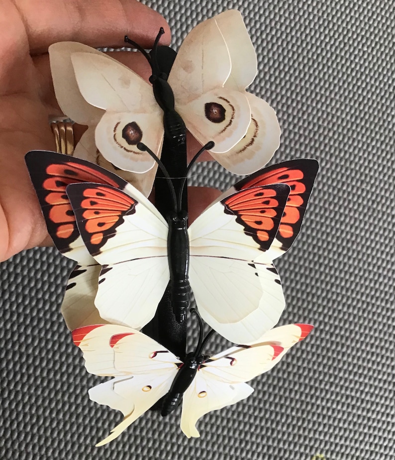 May include: Three butterfly hair clips with black bands. The top butterfly is white with brown markings. The middle butterfly is white with black and orange markings. The bottom butterfly is white with yellow markings.