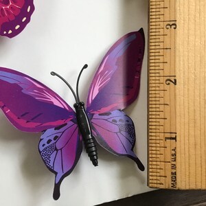May include: Purple butterfly wall decor with a black body and a ruler for size reference. The butterfly measures approximately 3 inches in length.