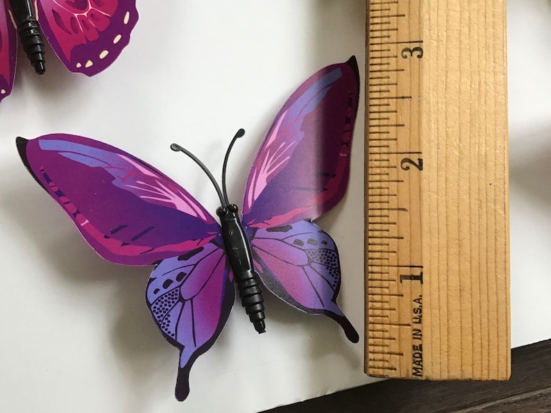 May include: Purple butterfly wall decor with a black body and a ruler for size reference. The butterfly measures approximately 3 inches in length.