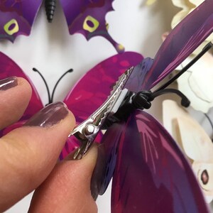 May include: A hand holding a silver butterfly clip with a black base. The clip is attached to a purple butterfly with iridescent wings.