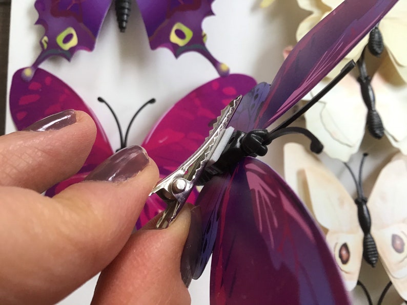 May include: A hand holding a silver butterfly clip with a black base. The clip is attached to a purple butterfly with iridescent wings.