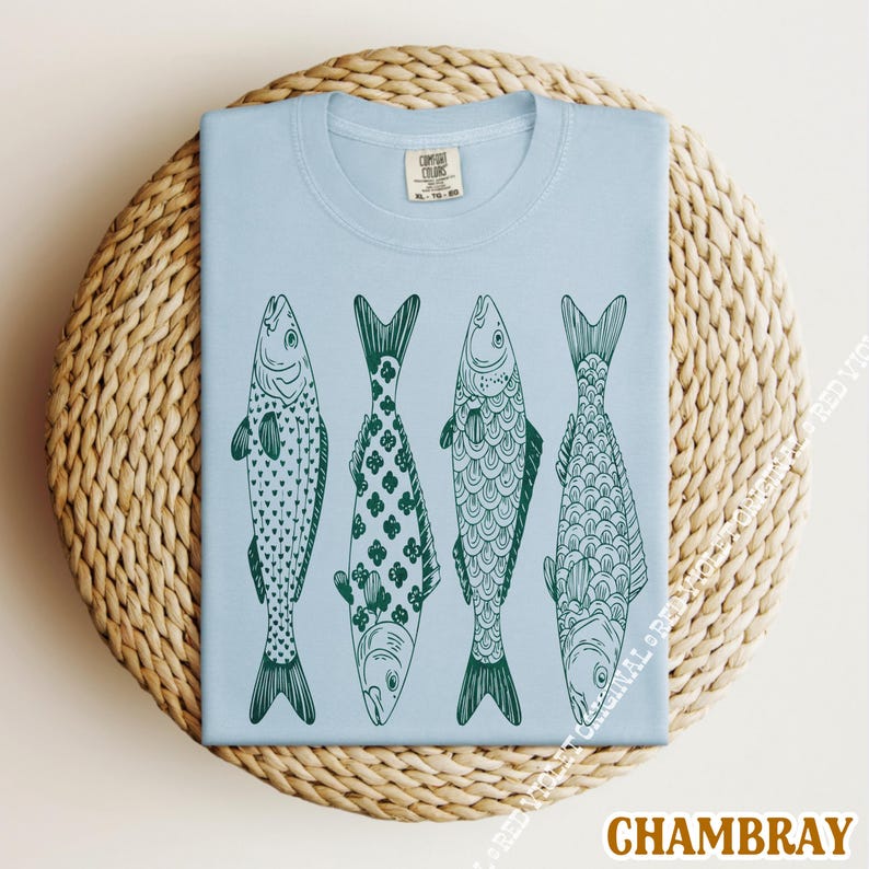 Op de afbeelding: Lichtblauw T-shirt met een ontwerp van vier gestileerde groene vissen, elk met een uniek patroon. Het shirt is opgevouwen op een ronde, geweven ondergrond. Het woord "CHAMBRAY" staat onderaan.