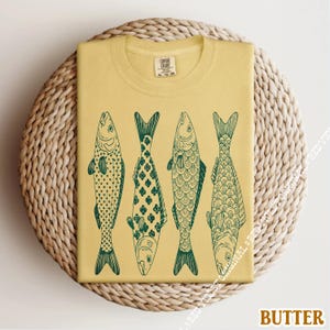 Op de afbeelding: Een gevouwen, boterkleurig t-shirt met een groene illustratie van vier vissen. Elke vis heeft een uniek schubbenpatroon en een gedetailleerd ontwerp. Het shirt ligt op een geweven, cirkelvormig oppervlak.