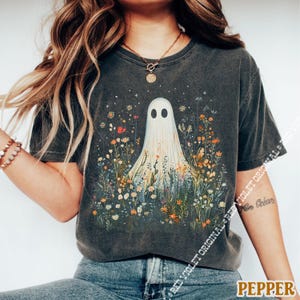 May include: Dark gray t-shirt featuring a whimsical ghost design amidst a field of colorful wildflowers. The ghost has two black oval eyes and a flowing white sheet. The word "PEPPER" is printed in a brown font at the bottom right.