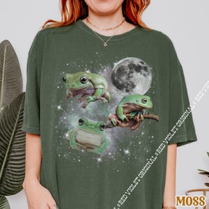 May include: A moss green t-shirt featuring a whimsical design of three green frogs against a backdrop of a starry galaxy and a full moon. The frogs are in various poses, adding a playful touch to the shirt's design.