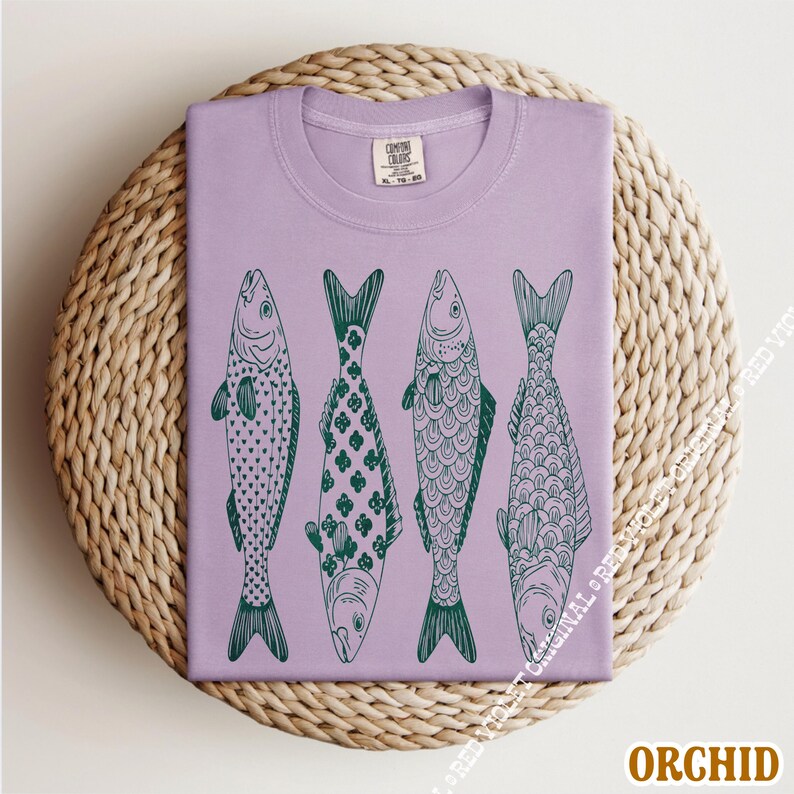 Op de afbeelding: Orchidee-kleurig T-shirt met vier gestileerde groene visillustraties. Elke vis heeft een uniek patroon, waaronder stippen, klavers en schubben. Het shirt is op een geweven, cirkelvormig oppervlak gevouwen. De tekst "ORCHID" staat rechtsonder.