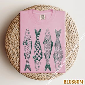 Op de afbeelding: Lichtroze T-shirt met vier gestileerde vissen in een rij, elk met een uniek donkergroen patroon. Het shirt is opgevouwen op een ronde, geweven ondergrond. Het woord "BLOSSOM" staat onderaan.