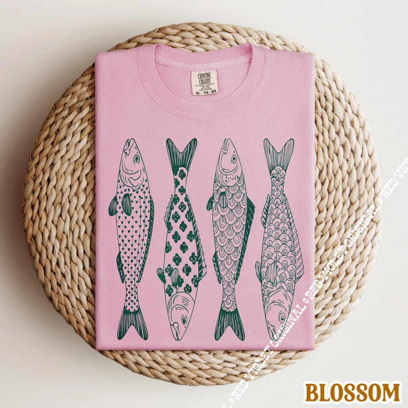 Op de afbeelding: Lichtroze T-shirt met vier gestileerde vissen in een rij, elk met een uniek donkergroen patroon. Het shirt is opgevouwen op een ronde, geweven ondergrond. Het woord "BLOSSOM" staat onderaan.