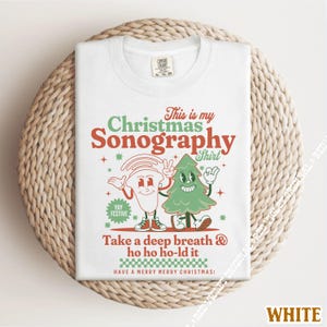 May include: White t-shirt with a Christmas-themed graphic. The design features a cartoon ultrasound image and a Christmas tree character with the text "This is my Christmas Sonography Shirt." Additional text includes "Take a deep breath & ho ho ho-ld it."