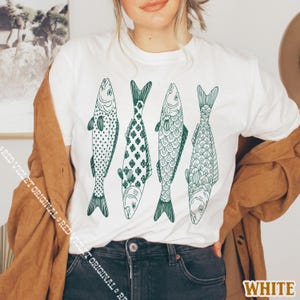 Op de afbeelding: Wit t-shirt met vier groene visillustraties. De vissen hebben verschillende patronen, waaronder harten, bloemmotieven en schubachtige details. Het woord "WHITE" is rechtsonder zichtbaar.