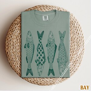 Op de afbeelding: Saliegroen t-shirt met vier gestileerde vissen op een rij, elk met een uniek donkergroen patroon. Het shirt wordt gepresenteerd op een ronde, geweven, lichtbruine ondergrond. Het woord "BAY" staat rechtsonder.