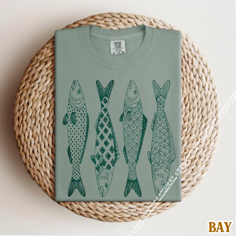 Op de afbeelding: Saliegroen t-shirt met vier gestileerde vissen op een rij, elk met een uniek donkergroen patroon. Het shirt wordt gepresenteerd op een ronde, geweven, lichtbruine ondergrond. Het woord "BAY" staat rechtsonder.