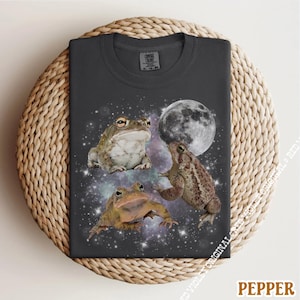 May include: Dark gray t-shirt featuring three frogs with a galaxy and moon design. The shirt has a crew neck and short sleeves. The image includes the text "PEPPER" in the bottom right corner.
