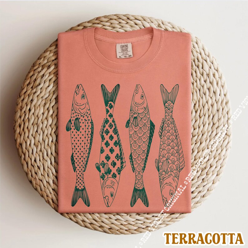 Op de afbeelding: Terracotta T-shirt met vier groene, gestileerde visontwerpen. Elke vis heeft een uniek patroon, met details zoals schubben en vinnen. Het shirt is opgevouwen op een ronde, geweven ondergrond. Onderaan staat 'TERRACOTTA'.