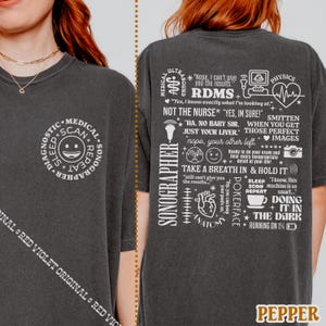 May include: Dark gray t-shirt with white text and graphics. The front features a circular design with "Diagnostic Scan Repeat" and a smiling face. The back has medical-themed phrases and images, including "Sonographer" and "RDMS".