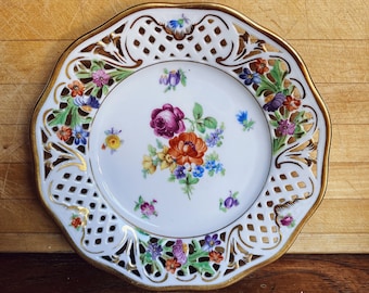 Peut inclure: Une assiette en porcelaine blanche avec un bord doré et un motif découpé complexe. L'assiette présente un motif floral avec des fleurs roses, orange, jaunes et bleues.