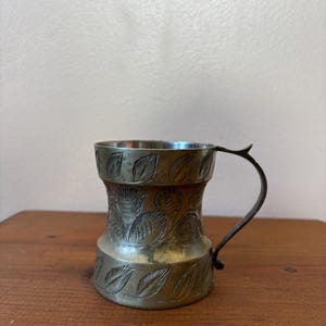 May include: A small, antique metal mug with a curved handle. The mug has an etched leaf design around the body. The metal has a tarnished, aged appearance, suggesting it is vintage. The mug sits on a wooden surface.