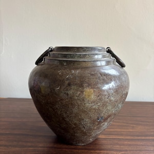 Puede incluir: Un jarrón de metal de color bronce vintage con dos asas de metal oscuro. El jarrón tiene un cuerpo redondeado y una superficie ligeramente texturizada. El jarrón está sobre una superficie de madera oscura.