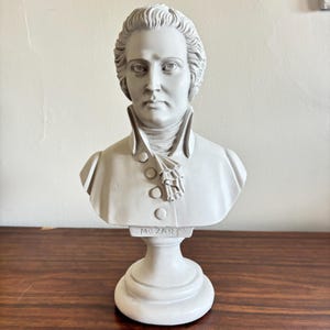 May include: A light gray bust of Mozart, featuring detailed facial features and a powdered wig. The bust is mounted on a pedestal base, with the name "MOZART" inscribed on the front. The sculpture is a decorative item.