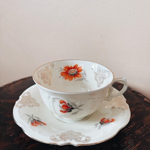 May include: A white teacup and saucer set with a floral design. The cup and saucer have a gold trim and feature orange and gray flowers.