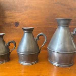 May include: Three pewter pitchers of varying sizes. The largest pitcher has a wide, rounded body and a handle. The two smaller pitchers have a more slender body and a handle.