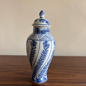 May include: A blue and white ceramic jar with a lid. The jar features a floral and leaf design in blue against a white background. The jar is approximately 6 inches tall.