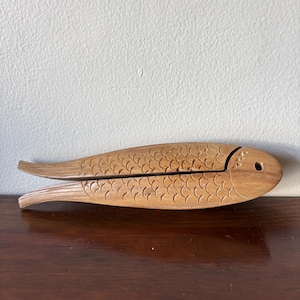 Puede incluir: Un cascanueces de madera con forma de pez. El cascanueces está tallado con detalles de escamas y tiene un diseño de bisagra. El pez es de color marrón claro y se encuentra sobre una superficie marrón oscuro.