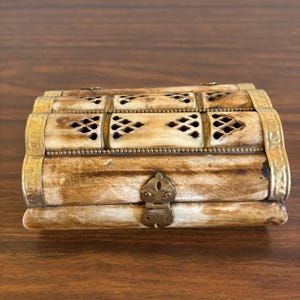 May include: A rectangular, antique-style box with a hinged lid. The box is made of light-colored wood or bone, accented with brass or gold-colored metal. Decorative cutouts and a clasp add detail to this vintage storage item.