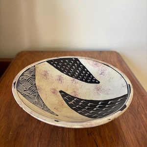 May include: A round ceramic bowl with a cream-colored interior and a dark brown rim. The bowl features black and white abstract designs, including curved lines and triangular shapes. The bowl is placed on a wooden surface.