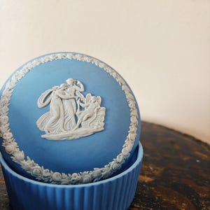 Puede incluir: Una caja de cerámica azul y blanca con un diseño en relieve de tres figuras y un borde floral.