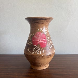 Może przedstawiać: Mały, brązowy ceramiczny wazon z rozszerzoną górą i podstawą. Wazon posiada malowaną różową różę i białe kwiatowe akcenty. Wazon jest eksponowany na drewnianej powierzchni na neutralnym tle.
