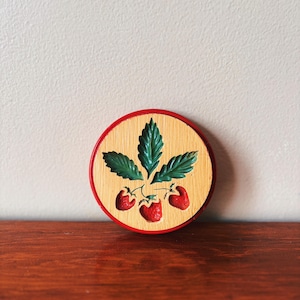 May include: A round wooden cookie cutter with a red rim. The cutter is shaped like a strawberry plant with three red strawberries and green leaves.